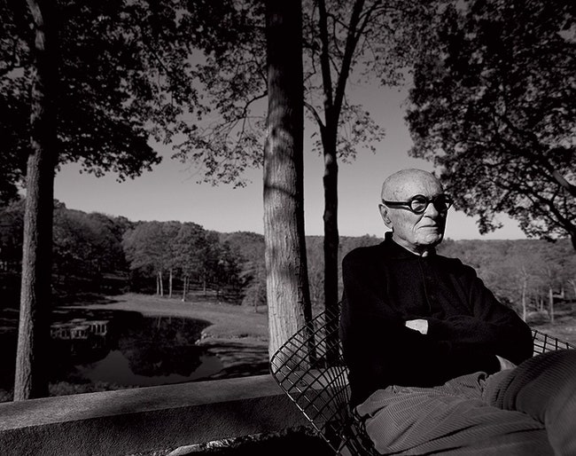 Филип Джонсон сидит в кресле по дизайну Гарри Бертойи в саду возле своего “Стеклянного дома” в НьюКанаане штат Коннектикут .