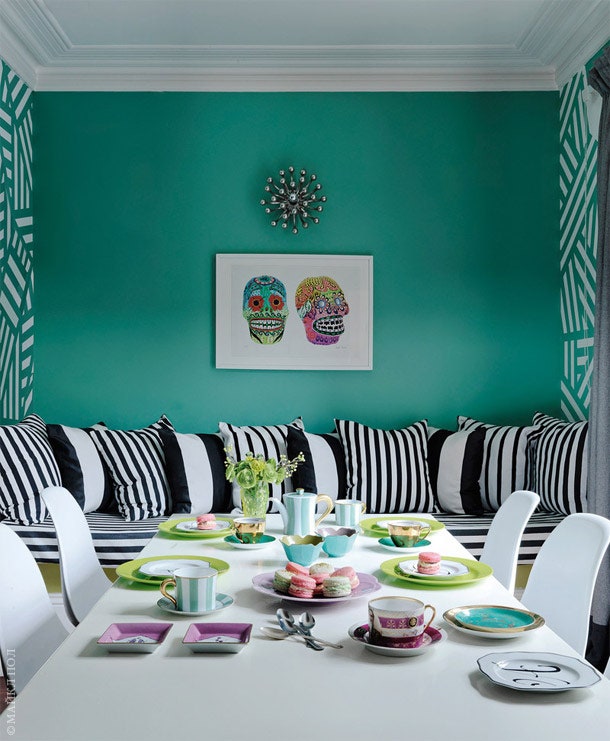 Обеденный стол на кухне сервирован к чаю. Одна из стен выкрашена бирюзовой краской — чернобелые подушки на кушетке...